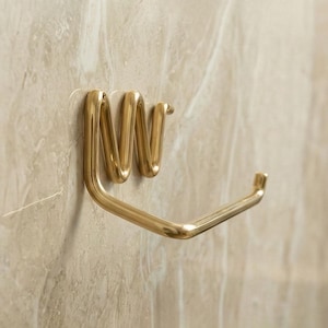 May include: A gold-colored, wall-mounted toilet paper holder. The holder has a unique, modern design with a coiled top section and a straight bar for holding the roll. It is mounted on a light beige, marble-like wall.