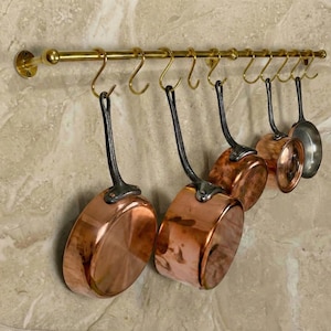 May include: A selection of copper saucepans suspended from a brass-coloured rail. The pans have dark handles and are of different sizes. The rail is mounted on a light-coloured, textured wall.