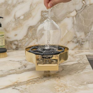May include: A gold-colored wine glass rinser with a black rubber insert is shown with water being poured into a wine glass. The rinser is mounted on a marble countertop. The wine glass is clear.