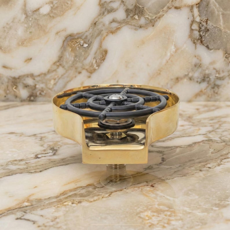 May include: A brass-colored beer tap with a black and gray wheel-shaped handle. The tap has a curved design and a threaded base for installation. The image is set against a marble background.