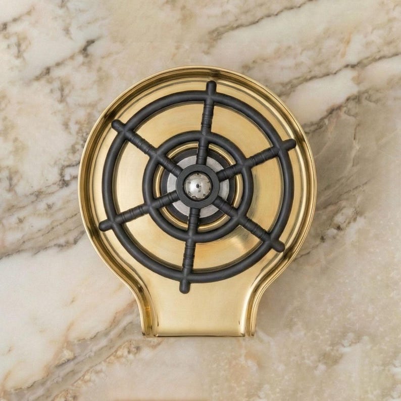 May include: A brass-colored soap dish with a black, ship's wheel-shaped insert. The dish has a rounded shape with a handle-like extension at the bottom. The background is a marble-like surface.
