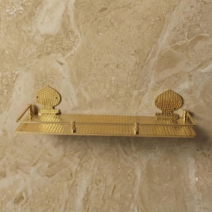 Peut inclure: Une étagère en métal doré avec une surface texturée et des accents décoratifs. L'étagère comporte deux supports ornés en forme de cœur et une petite barre à l'avant. L'étagère est fixée sur un mur beige et marron semblable à du marbre.