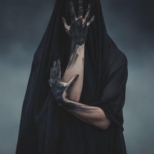 Könnte beinhalten: Eine Person in einem schwarzen Umhang, mit Armen und Händen, die mit dunkler Farbe bedeckt sind. Ein goldener Ring ist an einem Finger zu sehen. Der Hintergrund ist ein verschwommenes, dunkles Grau. Das Bild erzeugt ein Gefühl von Geheimnis und Dunkelheit.