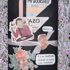 May include: A black envelope secured with a white ribbon and a wax seal, featuring a "MY BOOKSHELF" card, a Tazo tea bag, a pink pen, a sticker of a person reading, a floral sticker, and a note listing "Romance," "4.2 on Goodreads," "Friends to lovers," and "Dual timeline."