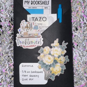 May include: A black envelope containing a My Bookshelf card, a Tazo tea bag, and a blue pen. It features a Booktrovert sticker, a floral sticker, and a white label with the text Romance, 3.8 on Goodreads, Fake Identity, and Dual POV printed on it.