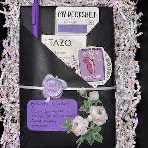 May include: A black wrapped book package featuring a purple pen, a Tazo tea bag, and stickers labeled My Bookshelf, Book On World Off, and Tell Me. A purple label lists Romantic Comedy, 3.8 on Goodreads, online to irl romance, found family, and gamers.