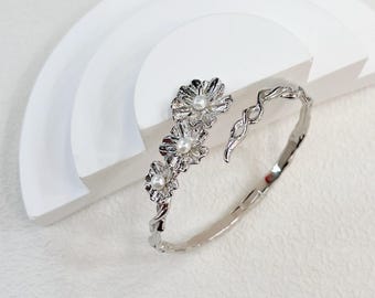 May include: A silver bangle bracelet with a floral design featuring three faux pearl accents. The bracelet is open and has a twisted metal design.