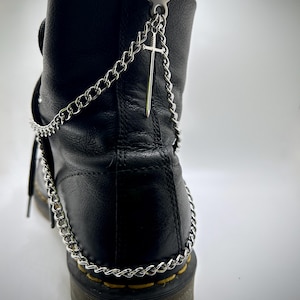 May include: A black leather boot adorned with a silver chain and a sword pendant. The chain encircles the heel, attaching to a metal ring at the boot's top. The sword charm dangles from the chain.