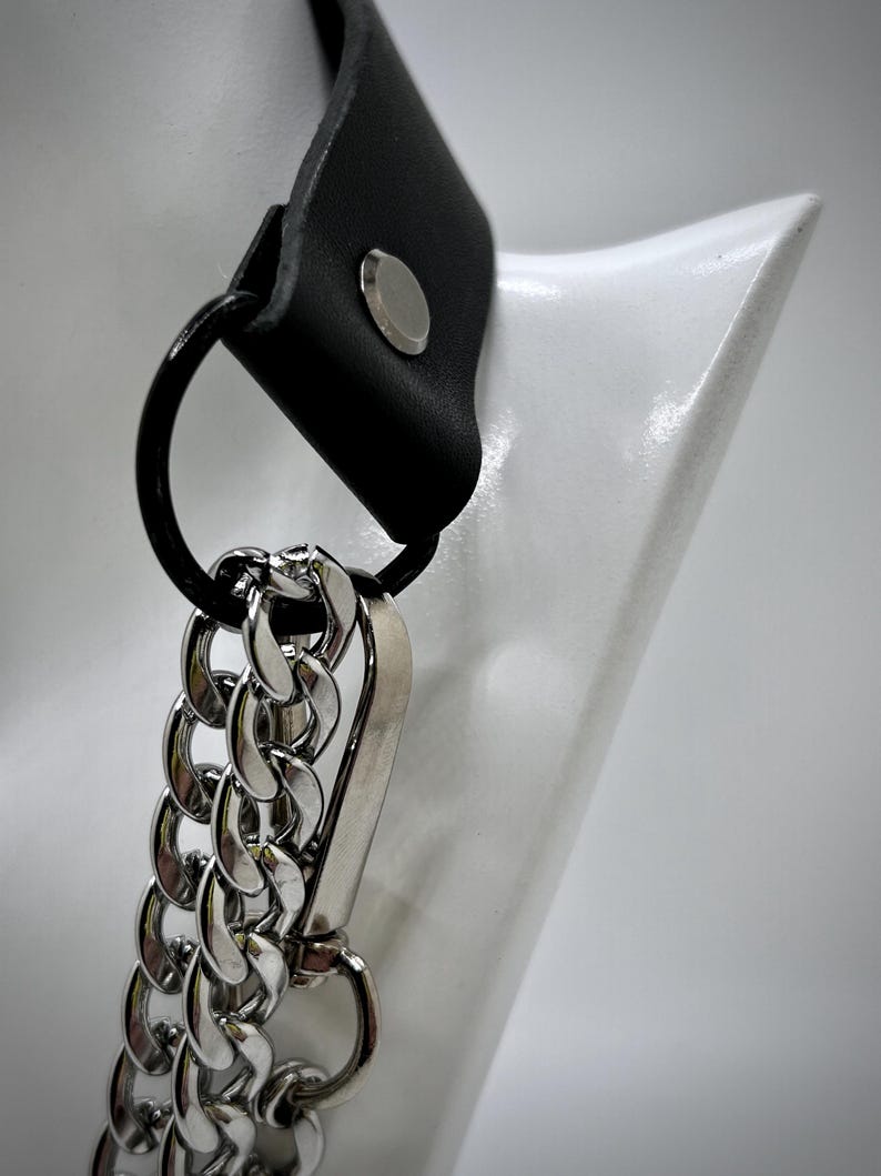 May include: A black leather choker with a silver chain and a silver clasp. The choker is on a white mannequin.