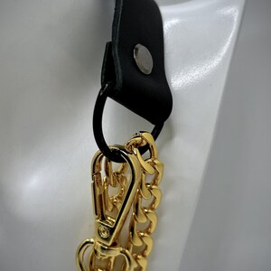 May include: A black leather strap with a gold metal chain and a gold metal clasp. The strap has a silver metal rivet.