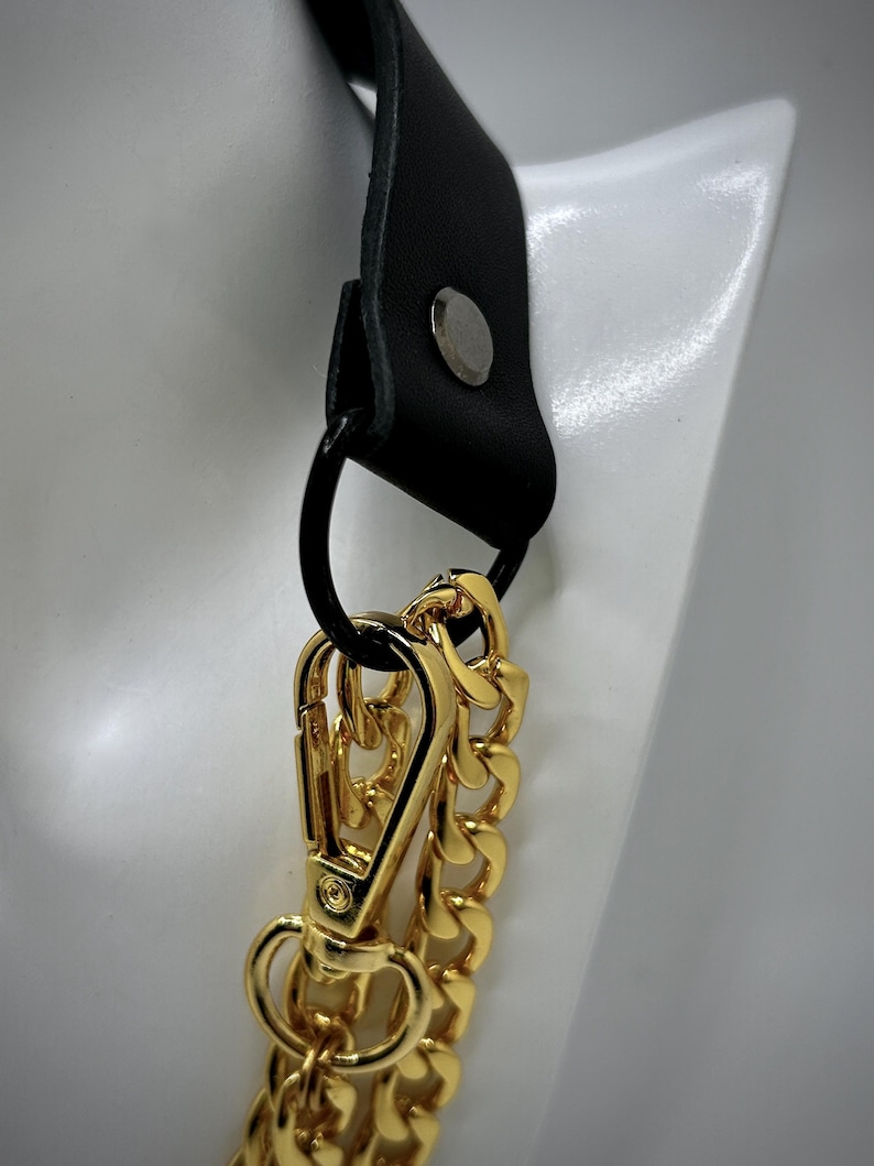 May include: A black leather strap with a gold metal chain and a gold metal clasp. The strap has a silver metal rivet.