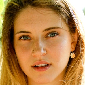 May include: A close-up portrait of a woman with long, blonde hair. She is wearing a gold necklace and small gold earrings. She has a nose ring and is looking directly at the camera.