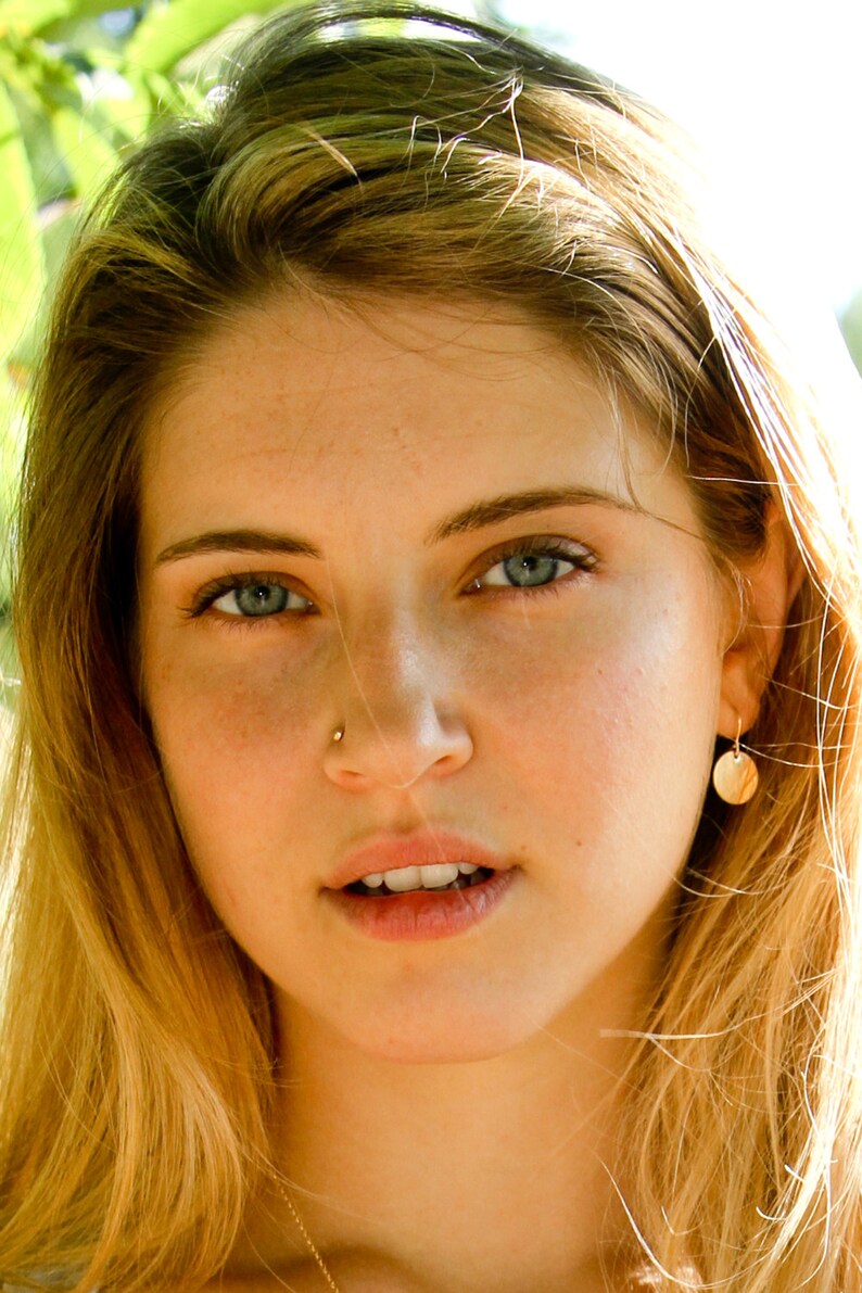 May include: A close-up portrait of a woman with long, blonde hair. She is wearing a gold necklace and small gold earrings. She has a nose ring and is looking directly at the camera.