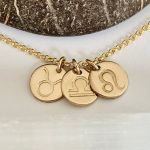 May include: Gold-colored necklace featuring three round pendants with zodiac symbols: Taurus, Libra, and Leo. The pendants are connected by a delicate chain. The background is a blurred, neutral color.