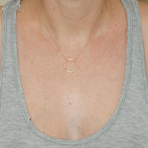 May include: A simple gold circle pendant necklace on a person wearing a grey tank top.