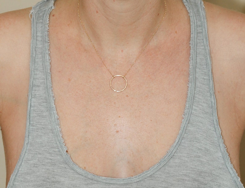 May include: A simple gold circle pendant necklace on a person wearing a grey tank top.