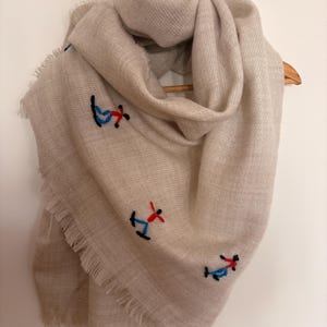 Puede incluir: Bufanda beige con figuras bordadas en rojo, azul y negro. La bufanda tiene un borde con flecos y está colgada de una percha de madera. La bufanda está hecha de un material suave y tejido.