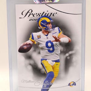 May include: A 2021 Prestige football card featuring Matthew Stafford in a Los Angeles Rams uniform. The card is encased in a protective sleeve. The card has a black and white background with the word "Prestige" at the top.