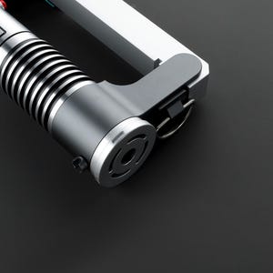 May include: A close-up of a silver lightsaber hilt with a black handle and a silver ring. The hilt has a black and silver design and a black button on the side.