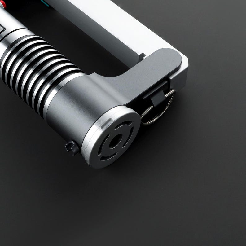 May include: A close-up of a silver lightsaber hilt with a black handle and a silver ring. The hilt has a black and silver design and a black button on the side.