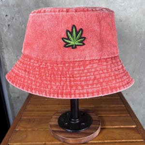May include: A red acid-wash bucket hat featuring a centered embroidered green cannabis leaf patch with a black outline. The hat has a wide, downward-sloping brim and a structured crown, showcasing a textured, faded fabric appearance throughout.