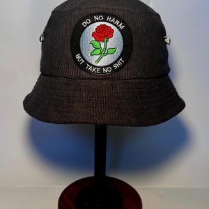 May include: Dark grey corduroy bucket hat with a circular patch featuring a red rose and the text "DO NO HARM BUT TAKE NO SHIT". The hat is displayed on a black stand with a wooden base.
