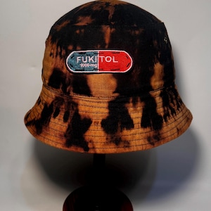 Puede incluir: Sombrero de pescador con efecto tie-dye marrón y negro. Cuenta con un parche con el texto "FUKITOL 1000 mg" en blanco, rojo y verde. El sombrero se exhibe sobre un soporte negro.