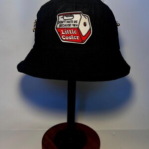 May include: Black corduroy bucket hat with a patch featuring a cooler design and the text "DON'T HATE ME BECAUSE I'M A Little Cooler." The hat has a small brim and is displayed on a black stand with a wooden base. Gold-colored embellishments are on the sides.