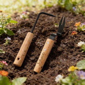 May include: Two gardening hand tools with wooden handles and black metal heads rest on soil. One tool has a curved head with a pointed end, and the other has a small fork-like head. The word "Johson" is engraved on the wooden handle.