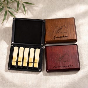 May include: Two rectangular wooden cases with magnetic closures and saxophone engravings. One case is open, revealing five light-coloured woodwind reeds held in a black interior. The other case is closed, featuring the text Saxophone and Everlasting Love.