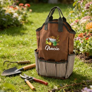 May include: A brown canvas garden tote bag with a striped bottom and multiple pockets. The bag has a green trim and handles. The word "Gracie" is printed on the front, along with a graphic of gardening tools. Gardening tools are in the foreground.