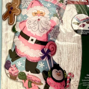 May include: A Bucilla Lollipop Santa felt stocking craft kit measuring 18 inches. The light blue stocking features a pink-clad Santa, a penguin, gingerbread man, cupcakes, and candy. The package includes stamped felt, sequins, and beads.