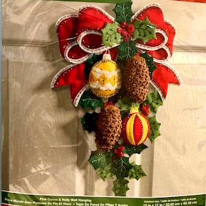 May include: A Bucilla Pine Cones & Holly wall hanging craft kit in original packaging. The kit features felt components including a large red bow, green holly leaves, red berries, two brown pine cones, and two patterned ornaments in yellow and red.