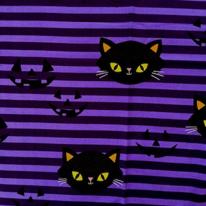 May include: Halloween-themed fabric with a repeating pattern of black cat faces and jack-o'-lanterns on a purple and black striped background. The cats have yellow eyes and ears, and white whiskers. The jack-o'-lanterns have a simple, smiling face.