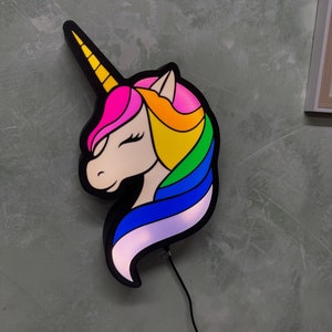 Könnte beinhalten: Eine farbenfrohe Einhorn-Wandleuchte mit schwarzer Umrandung. Das Einhorn hat ein gelbes Horn, eine pinke, orange, gelbe, grüne und blaue Mähne und ein weißes Gesicht. Das Licht ist an eine Steckdose angeschlossen.