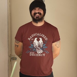 Puede incluir: Camiseta granate con las palabras "RADICALIZED by BASIC DECENCY" en un diseño circular. El diseño presenta dos palomas blancas y detalles florales azules. La camiseta tiene mangas cortas.