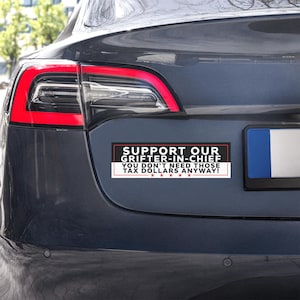May include: A rectangular bumper sticker with the text "SUPPORT OUR GRIFTER-IN-CHIEF YOU DON'T NEED THOSE TAX DOLLARS ANYWAY!" in black and red lettering. The sticker is affixed to the rear of a dark gray car.