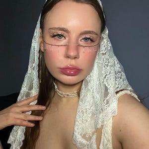 Op de afbeelding: Een vrouw draagt een witte kanten sluier en een delicaat zilveren ketting-oogaccessoire met bungelende druppelvormige bedels. Ze draagt ook een parelketting. De afbeelding heeft een zachte, romantische uitstraling.