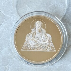 May include: A gold-colored coin featuring a detailed engraved image of a seated figure with a halo. The coin is encased in a clear plastic protective case. The figure is wearing a turban and robes, with one hand raised in a gesture of blessing.