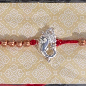May include: A red thread bracelet featuring a central silver-toned Ganesha charm flanked by metallic copper-colored beads. The bracelet is mounted on a rectangular card decorated with a light tan geometric pattern and illustrations of hands holding lit diyas.