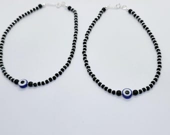 May include: Two beaded anklets with black beads and silver accents. Each anklet features a blue and white evil eye charm. The anklets are displayed on a white background.