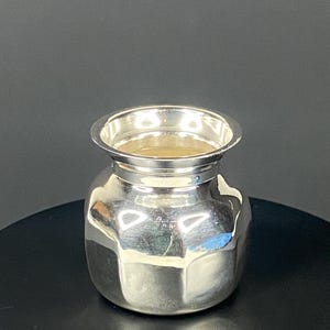 May include: A small, silver-colored vase with a wide, open top and a rounded body. The vase has a faceted design, giving it a unique, geometric appearance. The interior of the vase appears to be a light tan color. The vase is sitting on a black surface.