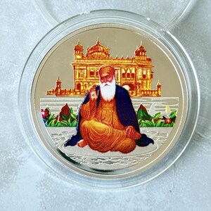 May include: A silver coin featuring a seated figure with a white beard and a turban, wearing orange and blue robes. Behind the figure is a detailed depiction of a golden building. The coin is encased in a clear plastic holder.