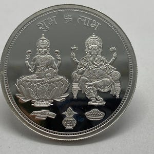 May include: A silver coin featuring embossed images of Hindu deities. The coin has the words "Shubh Labh" in Devanagari script at the top. The coin's design includes images of Lakshmi and Ganesha, symbols of prosperity.