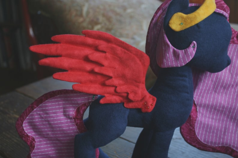 Op de afbeelding: Een pluche paard met een rode viltvleugel, een roze en wit gestreept stoffen lichaam en een blauwe viltkop met een gele vilt hoorn.