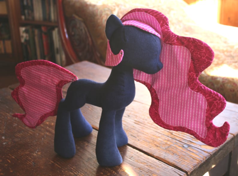 Op de afbeelding: Een pluche speelgoedpaard met een donkerblauw lichaam en een roze-wit gestreepte manen en staart. Het paard heeft een roze-wit gestreepte manen en staart.