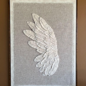 Op de afbeelding: Een witte vleugel sculptuur van stof, ingelijst in een donker houten frame. De vleugel is gemaakt van veel kleine, gelaagde stukken stof, waardoor een textuur en een driedimensionaal effect ontstaat.