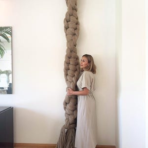 May include: A large, beige-colored, braided rope art piece, mounted on a wall. The rope is thick and textured, with a decorative black crown at the top. The rope extends to the floor, with a frayed end. The art piece is displayed in a well-lit room.