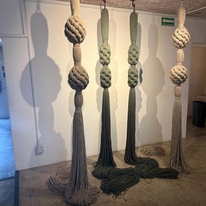 May include: Hanging decorative art pieces. The pieces are made of thick, textured rope in shades of beige and olive green. Each piece has a knotted design and a long tassel at the bottom. The art is suspended from the ceiling.