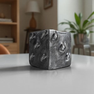 May include: A large, gray, metal dice with a textured surface. The dice is sitting on a white table.